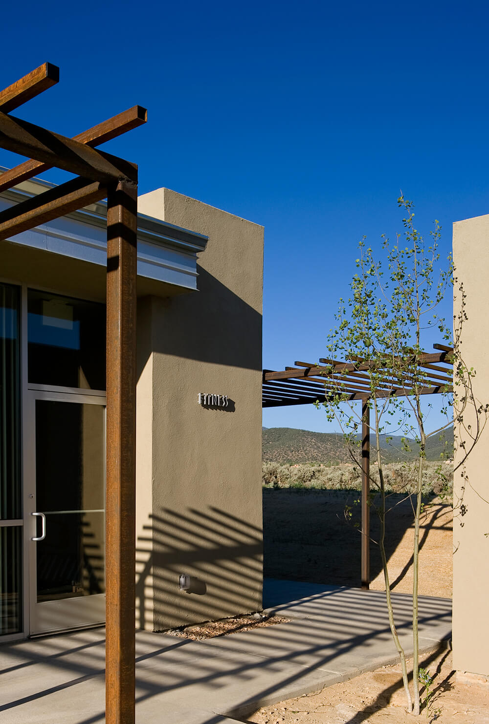 Steel pergola casting shadows across a community center walkway under a deep blue Taos sky
